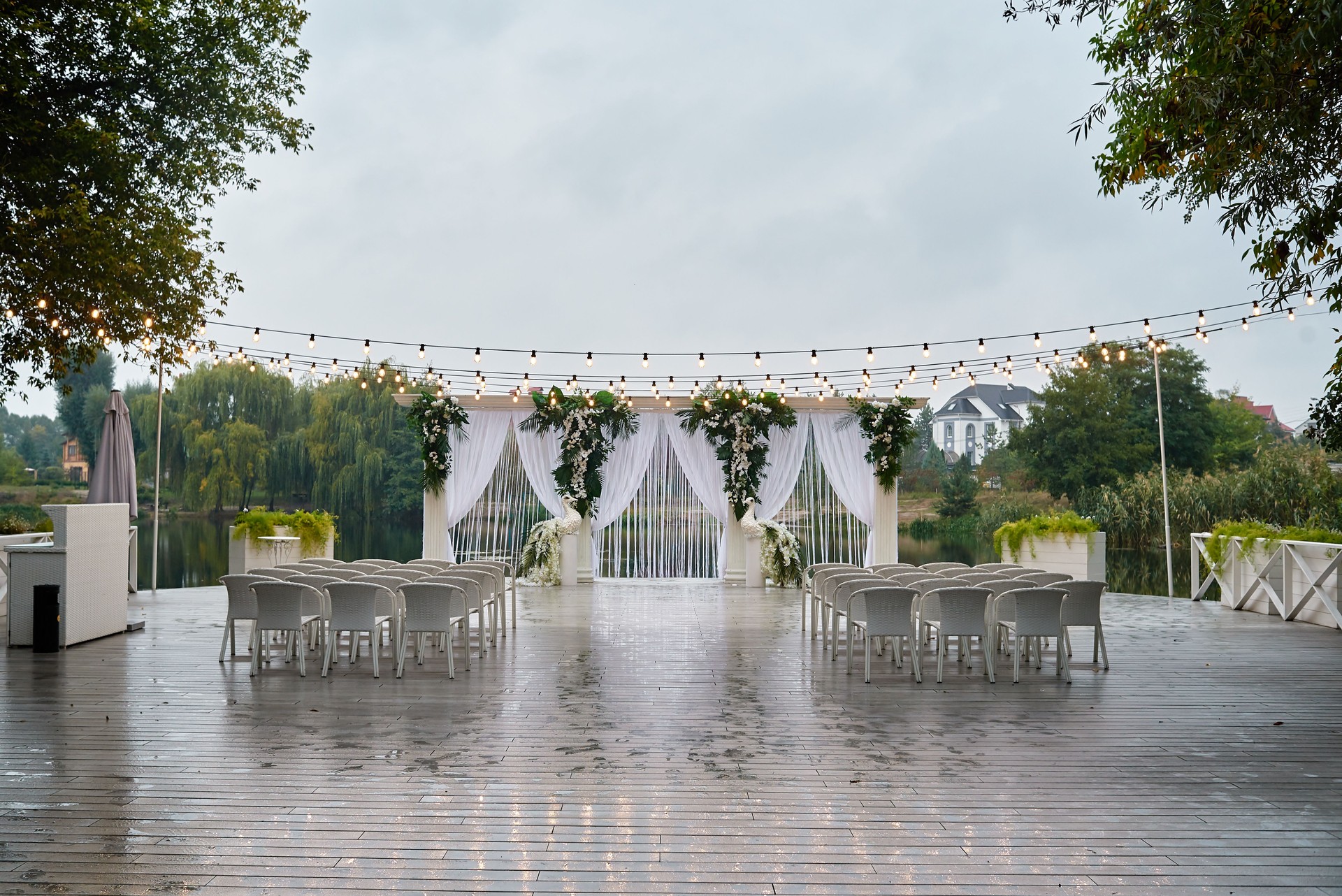 Place for wedding ceremony with wedding arch decorated with palm leaves, orchid flowers and floral peacocks, bulbs garland and white chairs outdoors, copy space